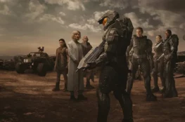 Grupa postaci z serialu "Halo" w futurystycznym otoczeniu. W centrum zdjęcia stoi żołnierz w zbroi, trzymający dużą broń. Po jego prawej stronie znajduje się kilka osób ubranych w wojskowe uniformy. W tle widoczny jest pojazd terenowy, a nad nimi pochmurne niebo.