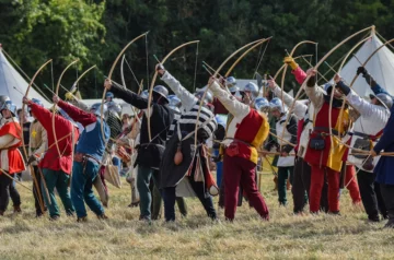 Na zdjęciu widać grupę osób ubranych w średniowieczne stroje, uczestniczących w rekonstrukcji historycznej związanej z łucznictwem. Osoby te trzymają łuki i są gotowe do oddania strzału. W tle widać namioty i drzewa.