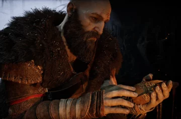 Kratos z poważnym wyrazem twarzy trzyma w dłoniach zdobiony amulet, pogrążony w głębokiej refleksji.