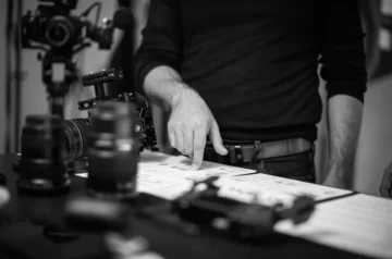 Czarno-białe zdjęcie przedstawia reżysera wskazującego na storyboardy. W tle znajduje się sprzęt filmowy, w tym aparat i obiektywy. Scena sugeruje pracę nad planem zdjęciowym lub organizację produkcji