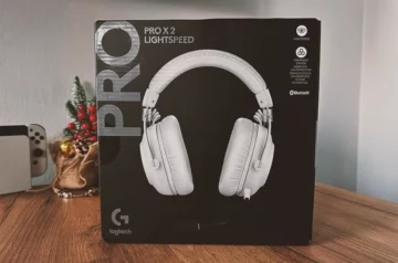 Opakowanie słuchawek Logitech G PRO X 2 LIGHTSPEED w wersji białej, ustawione na drewnianym stole. W tle widoczna świąteczna dekoracja oraz konsola Nintendo Switch OLED w wersji białej.