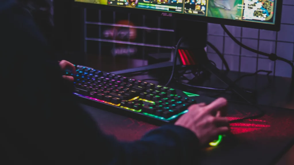 Zbliżenie na gamingową klawiaturę z kolorowym podświetleniem RGB w ciemnym pomieszczeniu. Widoczne są dłonie użytkownika podczas gry, a w tle monitor z wyświetloną grą. Klawiatura ma wyraźne, wielokolorowe podświetlenie klawiszy w odcieniach fioletowych, niebieskich, zielonych i żółtych, co tworzy charakterystyczną dla graczy atmosferę.