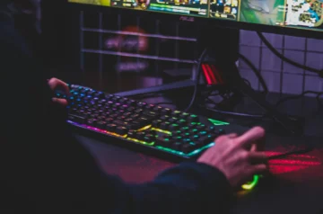 Zbliżenie na gamingową klawiaturę z kolorowym podświetleniem RGB w ciemnym pomieszczeniu. Widoczne są dłonie użytkownika podczas gry, a w tle monitor z wyświetloną grą. Klawiatura ma wyraźne, wielokolorowe podświetlenie klawiszy w odcieniach fioletowych, niebieskich, zielonych i żółtych, co tworzy charakterystyczną dla graczy atmosferę.