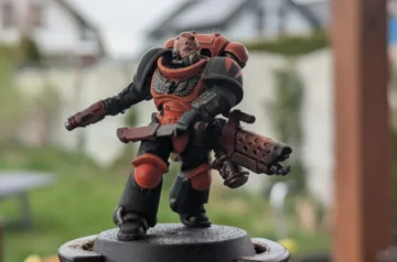 Pomalowana figurka kosmicznego marine’a z gry bitewnej Warhammer 40,000 stoi na podstawce modelarskiej na zewnątrz, na tle rozmytego ogrodu i budynków. Figurka ma czerwono-czarny pancerz, odsłoniętą głowę z brodą i bliznami, w lewej ręce trzyma dużą broń palną z otworami chłodzącymi, a w prawej miecz. Na piersi widoczny jest srebrny orzeł. Tło jest rozmyte, dzięki czemu figurka jest wyraźnie widoczna na pierwszym planie.