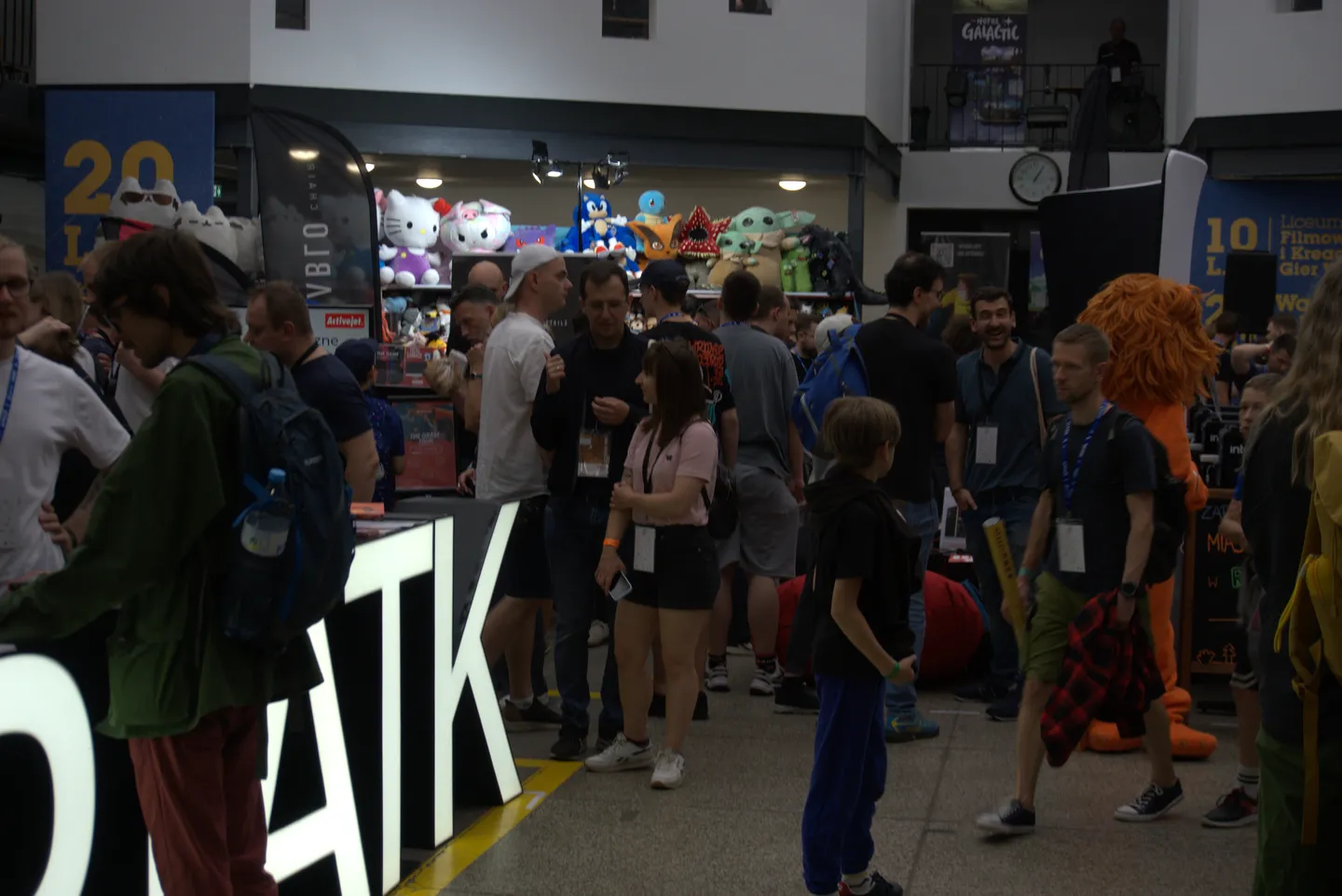 Tłum uczestnik&oacute;w Pixel Heaven 2025 w strefie targowej, otoczonej stoiskami z gadżetami, pluszakami i popkulturowymi maskotkami. W tle widoczne są duże postacie, takie jak Hello Kitty i Sonic, a atmosfera wydarzenia sprzyja spotkaniom i rozmowom fan&oacute;w gier oraz retro kultury