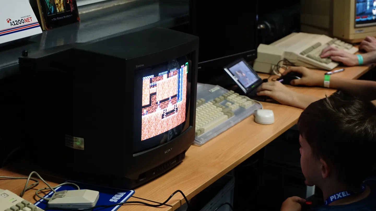Uczestnik festiwalu gra na klasycznym komputerze Amiga, korzystając z monitora CRT Sony. Na ekranie widoczna jest kultowa gra retro, a obok znajduje się myszka i klawiatura w stylu lat 90