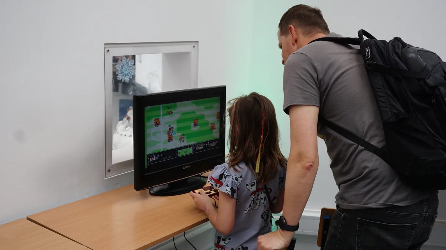 Dziewczynka gra w kolorową grę zręcznościową na retro konsoli pod czujnym okiem dorosłego. W tle widoczny telewizor CRT i minimalistyczne stanowisko do gier.