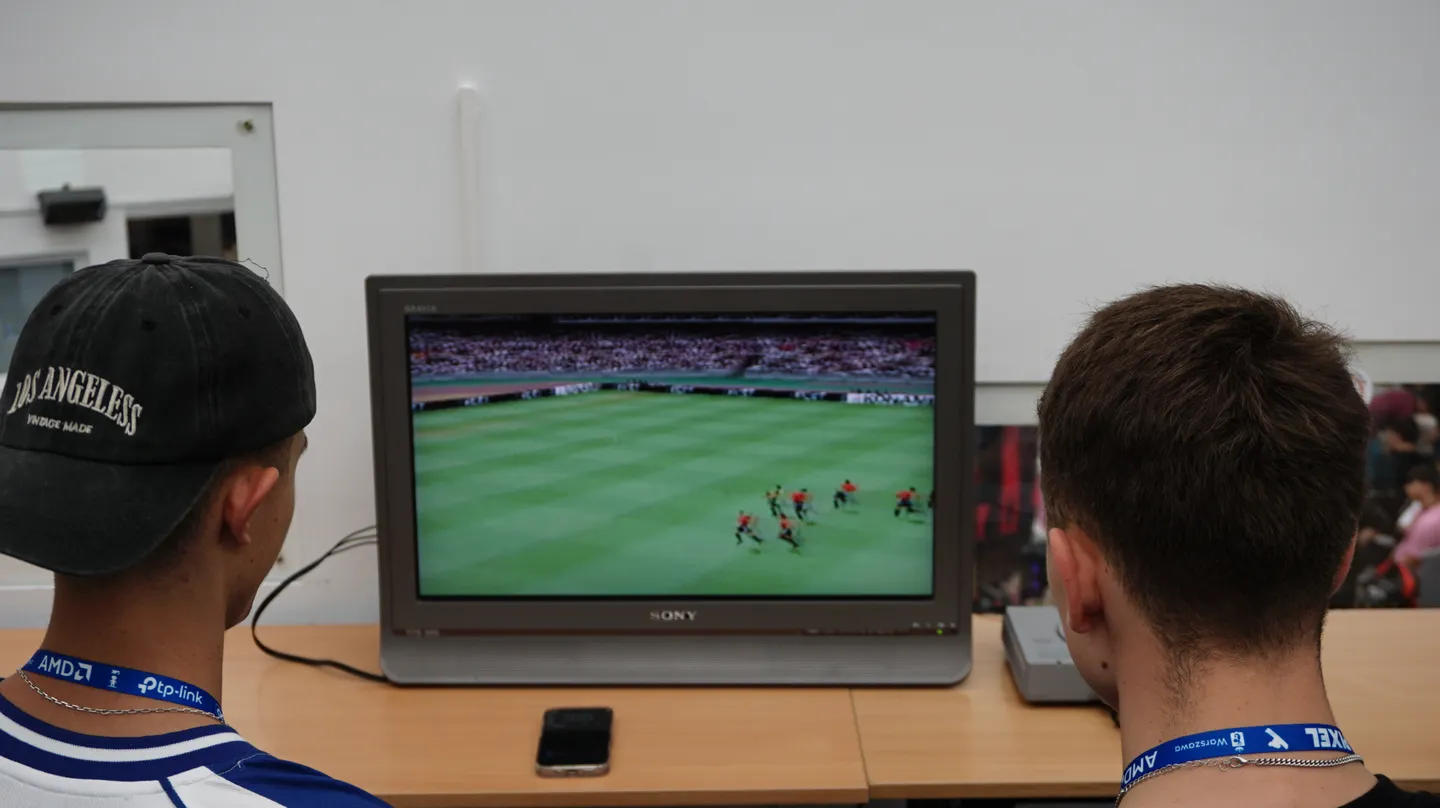 Dw&oacute;ch uczestnik&oacute;w gra w klasyczną grę piłkarską na konsoli retro, korzystając z telewizora CRT Sony. Atmosfera przypomina dawne czasy salon&oacute;w gier i domowych rozgrywek z lat 90.