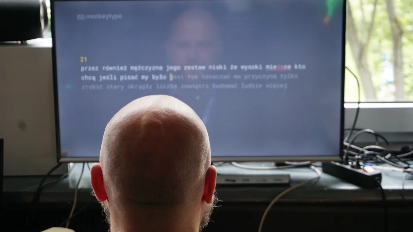 Mężczyzna bierze udział w konkursie szybkiego pisania na klawiaturze przy dużym monitorze, skupiając się na wyświetlonych zadaniach tekstowych.