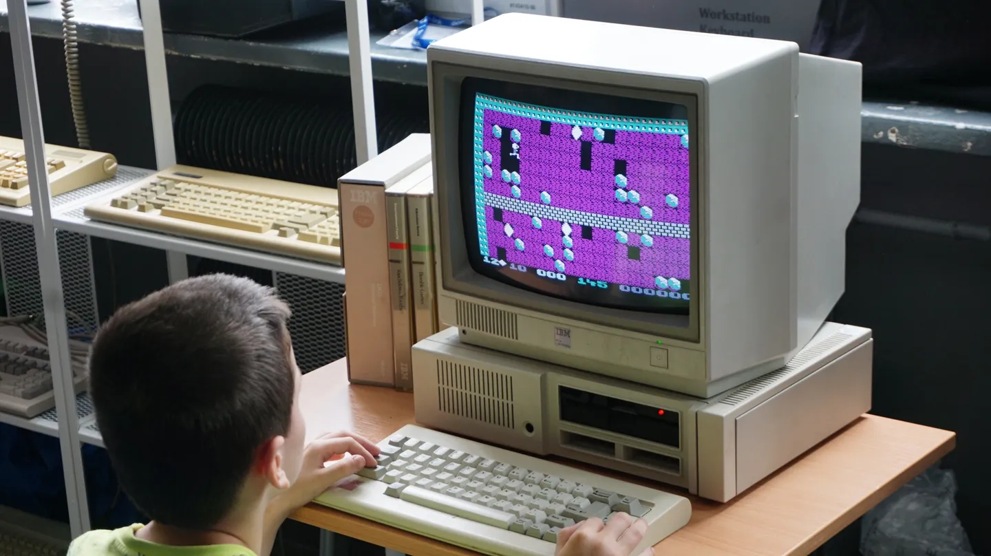 Chłopiec gra w klasyczną grę komputerową na starym komputerze IBM z oryginalnym monitorem i klawiaturą, otoczony retro sprzętem i dyskietkami.