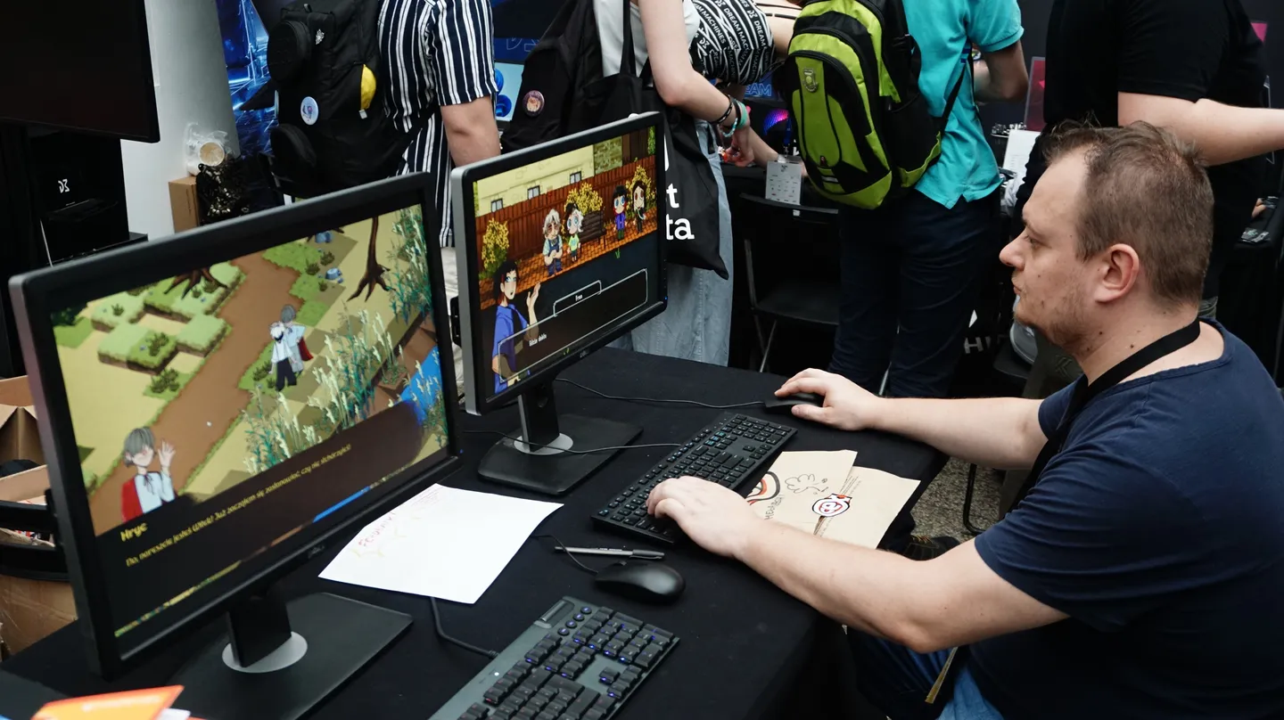 Uczestnik festiwalu testuje grę indie przy stanowisku z dwoma monitorami, korzystając z klawiatury i myszy, a w tle widać zwiedzających