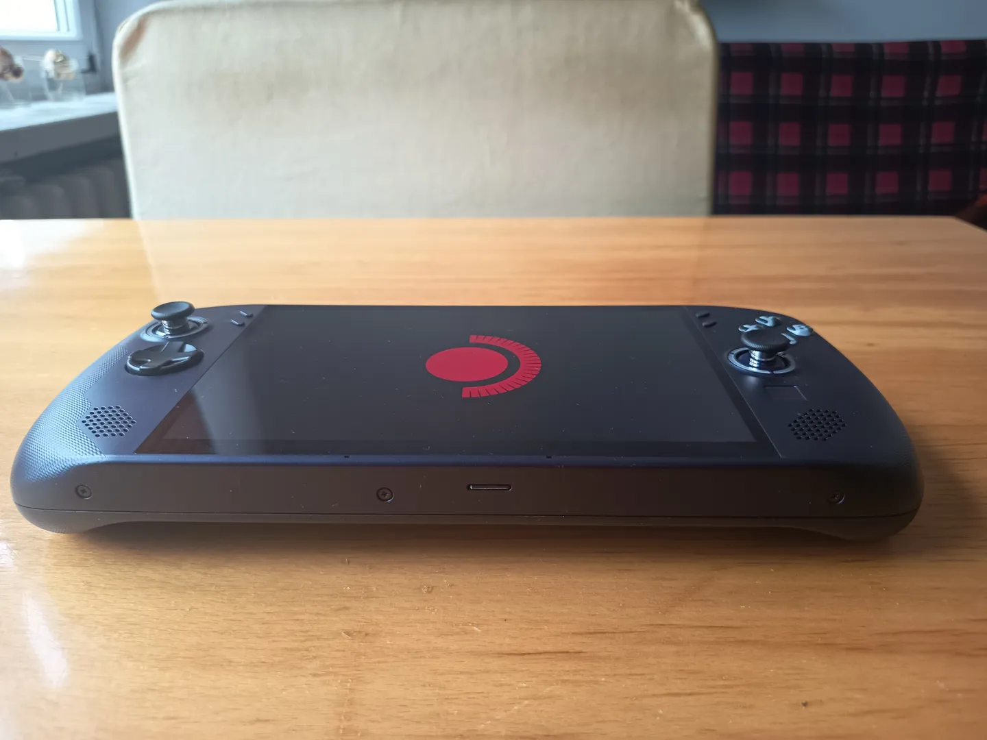 Widok z g&oacute;ry na przenośną konsolę do gier leżącą na stole, z wyświetlaczem w centrum i widocznymi głośnikami oraz gałkami analogowymi po bokach.