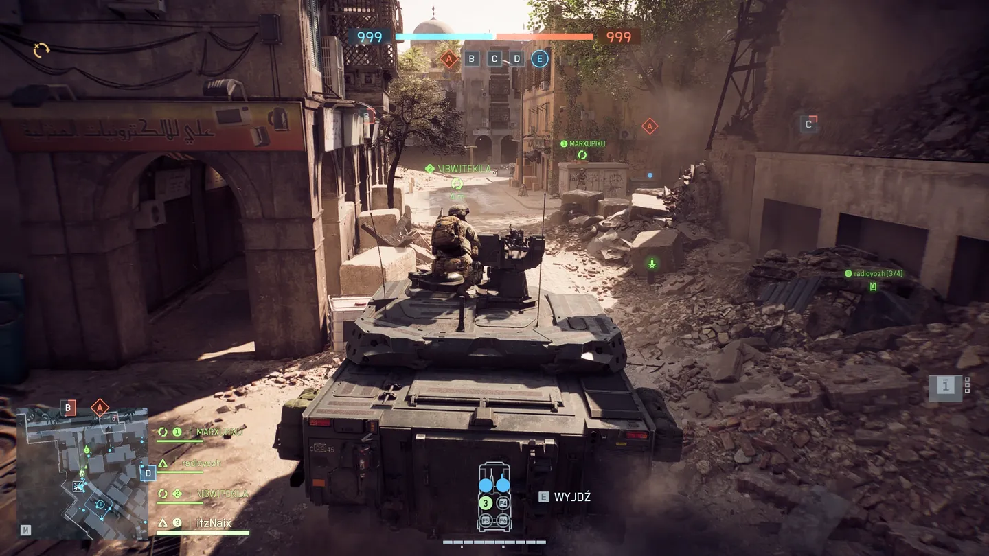 Widok zza pojazdu opancerzonego poruszającego się przez wąską uliczkę miasta, z żołnierzem obsługującym karabin maszynowy na dachu, otoczenie zniszczone po walkach; w lewym dolnym rogu minimapa i status drużyny.