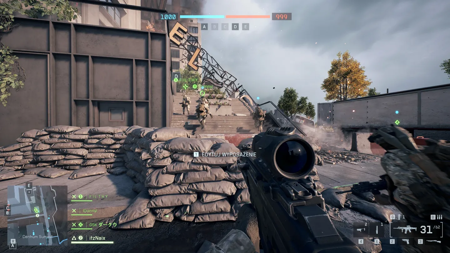 Widok z perspektywy gracza ukrytego za workami z piaskiem na placu budowy, celującego w kierunku schod&oacute;w prowadzących do wejścia budynku, po kt&oacute;rym biegną członkowie drużyny; w lewym dolnym rogu widoczna minimapa.