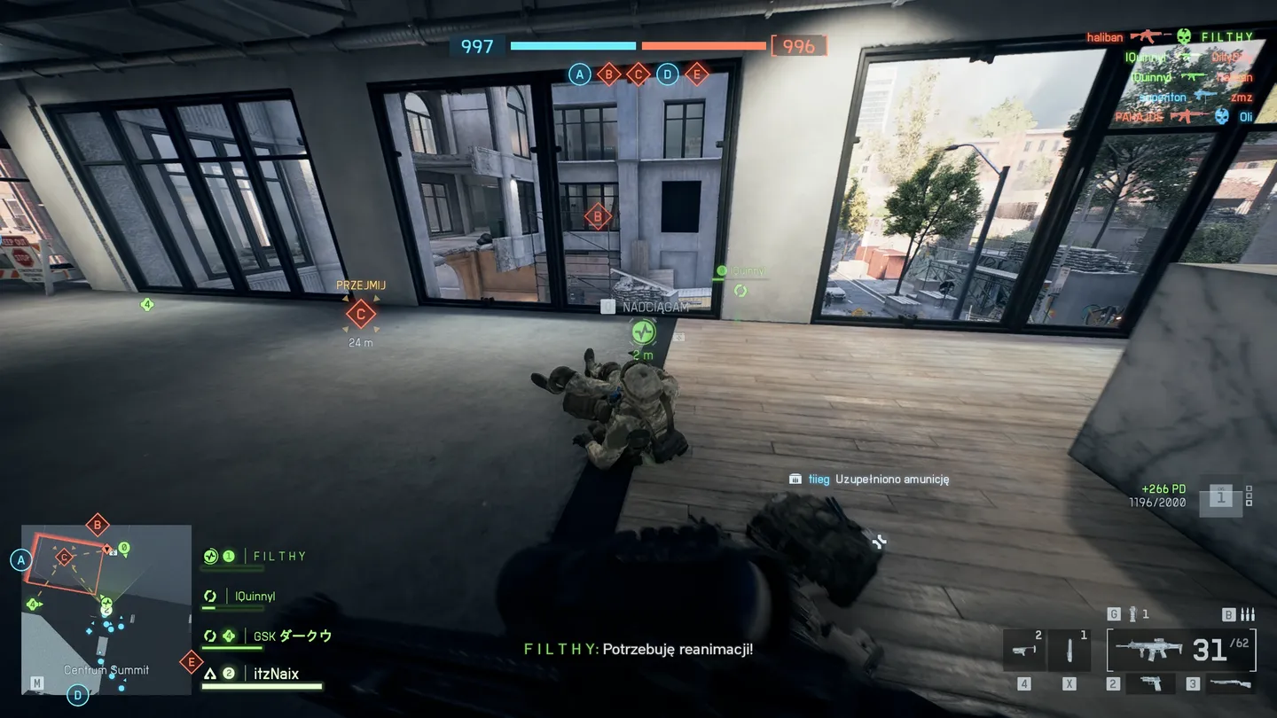 Wnętrze nowoczesnego budynku, perspektywa gracza na pomieszczenie z dużymi oknami i widokiem na zewnątrz; na środku leży powalony żołnierz, kt&oacute;remu pomaga sojusznik, a na ekranie widoczna minimapa oraz statusy drużyny i punkt&oacute;w kontrolnych.