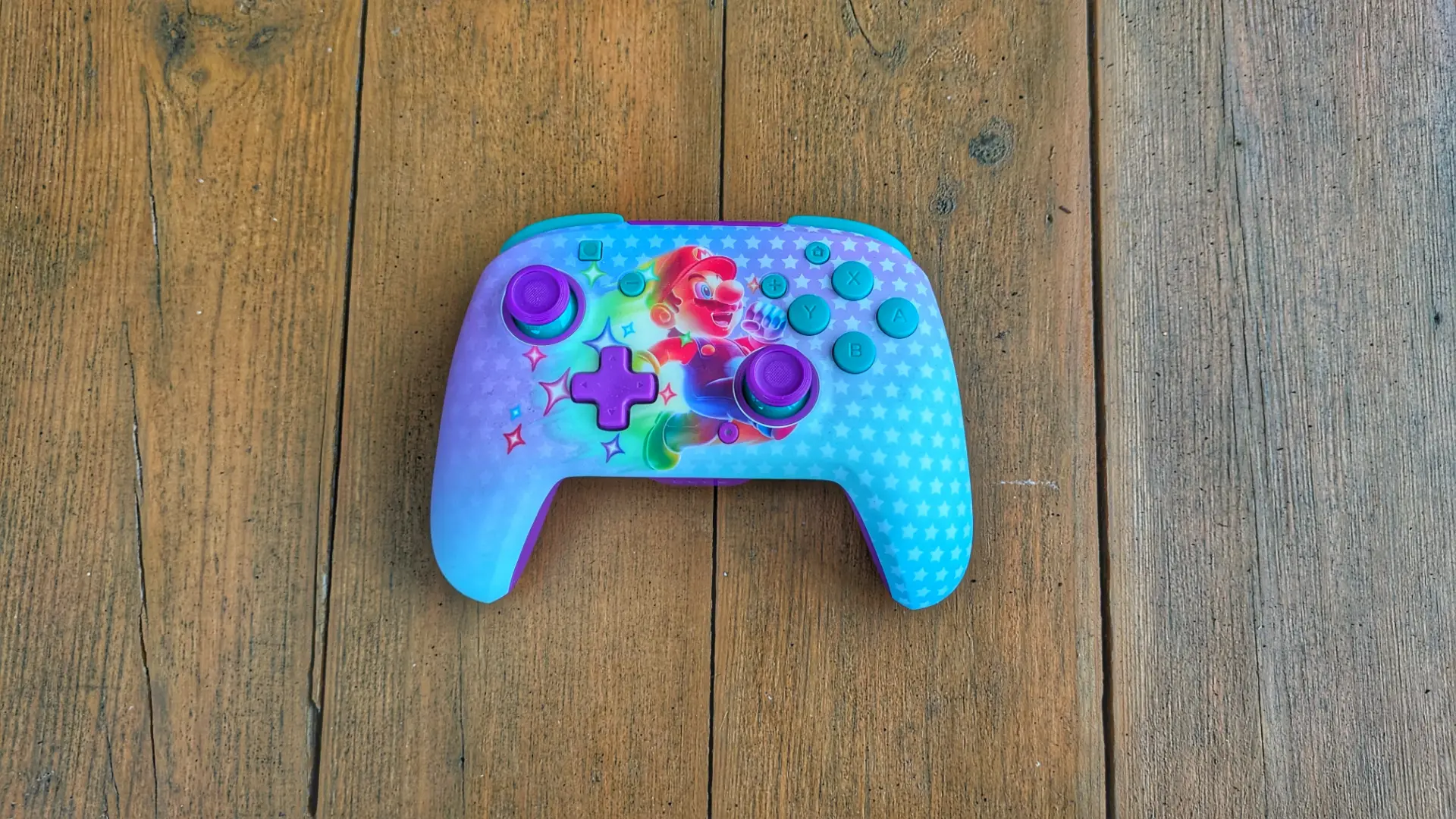Na zdjęciu znajduje się kolorowy kontroler do gier Turtle Beach Rematch Wireless Invincible Mario z motywem Mario, leżący na drewnianym stole.