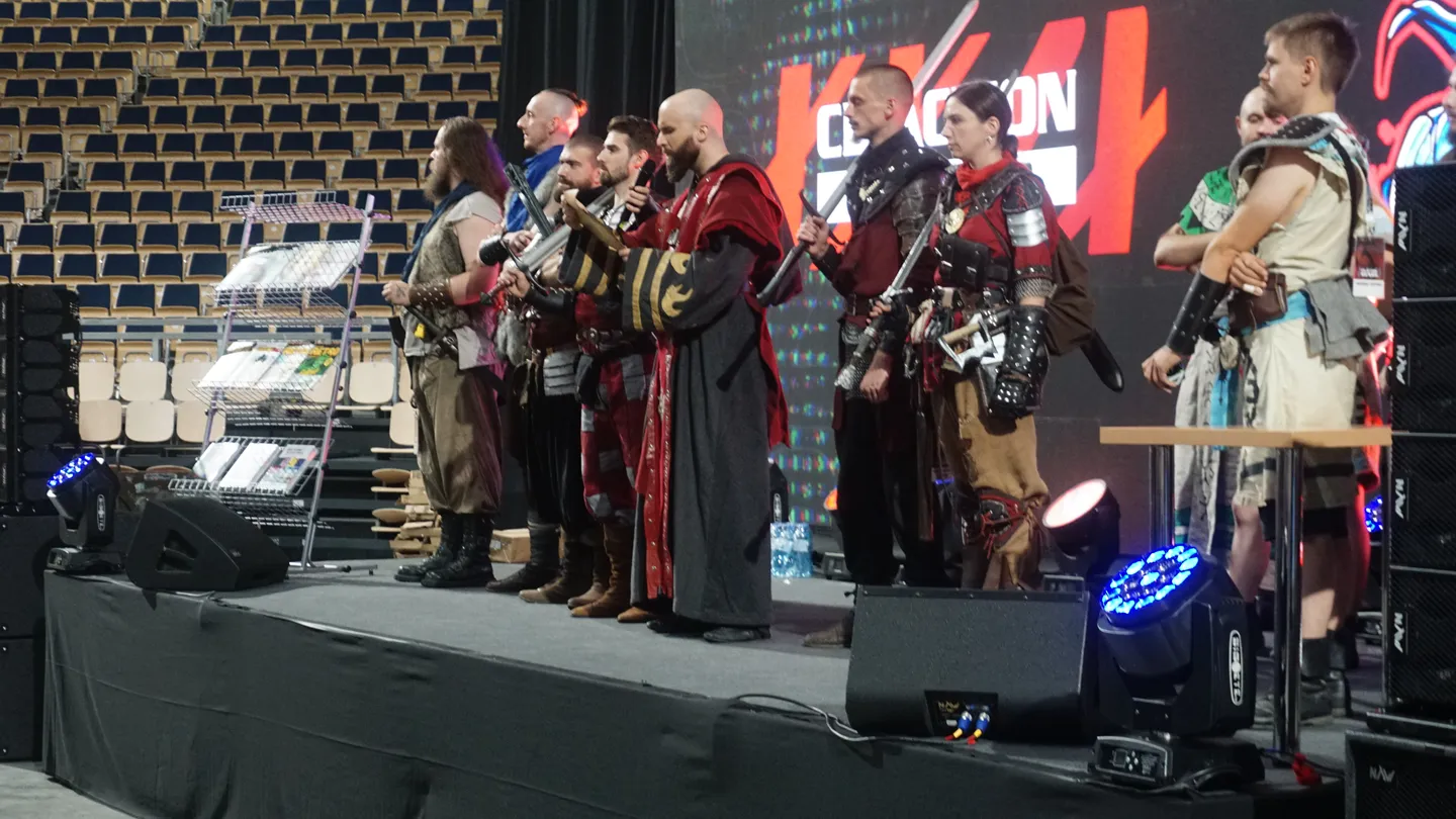 Cosplayerzy prezentują stroje inspirowane grami RPG na scenie CD-Action EXPO 2025. Wyraziste kostiumy, rekwizyty oraz pasja fan&oacute;w podkreślają kreatywność polskiej społeczności gamingowej. Scena na tle pustej widowni pokazuje przygotowania do konkursu cosplay.