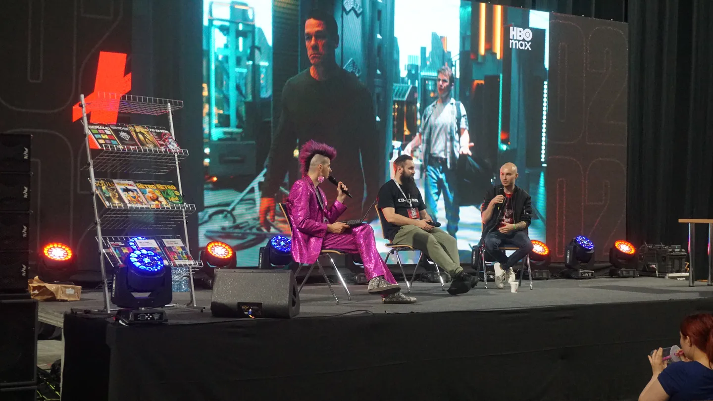 Panel dyskusyjny na scenie CD-Action EXPO 2025 &ndash; prowadzący w jaskrawo r&oacute;żowym garniturze rozmawiają z gośćmi na tle ekranu HBO Max, obok stojak z komiksami. Wyjątkowa atmosfera konwentu gamingowego w centrum wydarzeń branży gier.