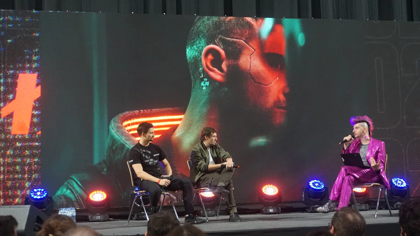 Wywiad z tw&oacute;rcami gier podczas CD-Action EXPO 2025. Host eventu w błyszczącym r&oacute;żowym garniturze prowadzi rozmowę z zaproszonymi ekspertami branży, na tle ekranu z motywem cyberpunkowym. Gamingowa konferencja w Polsce skupia fan&oacute;w i deweloper&oacute;w.