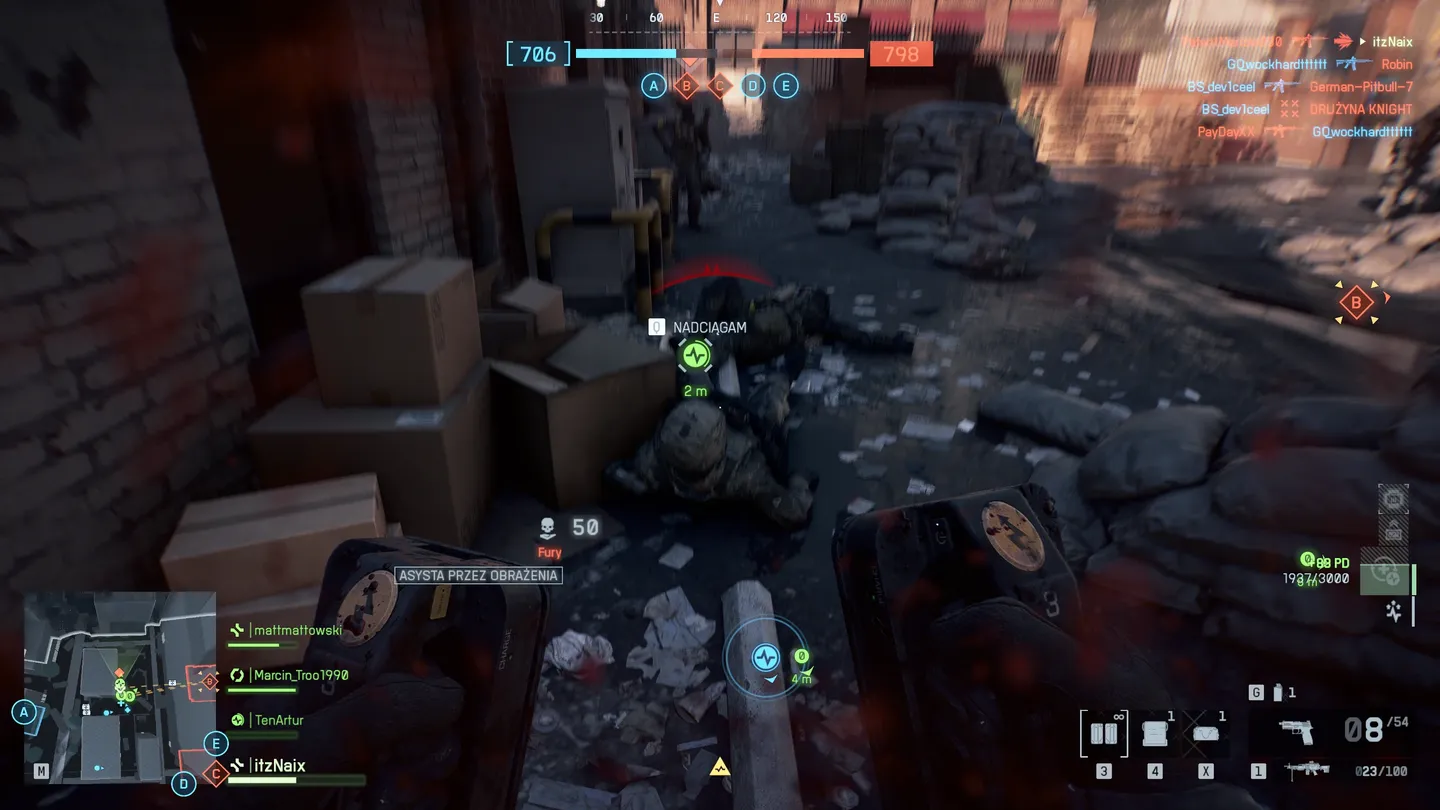 Widok z perspektywy pierwszej osoby podczas reanimacji sojusznika w ciasnej, zrujnowanej uliczce pełnej pudeł i work&oacute;w; na ekranie widać interfejs reanimacji, mapę, statystyki oraz niebieskie i czerwone wskaźniki kontrolowanych punkt&oacute;w.