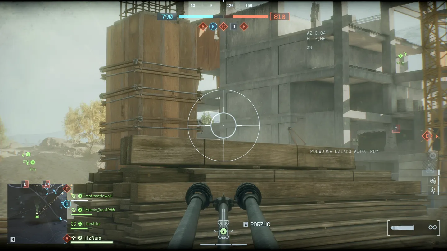 Widok z wnętrza pojazdu z podw&oacute;jnym działkiem automatycznym, skierowanym na budowę z betonowymi ścianami i drewnianymi belkami; na ekranie widoczny jest celownik, informacje o broni oraz minimapa i status drużyny.