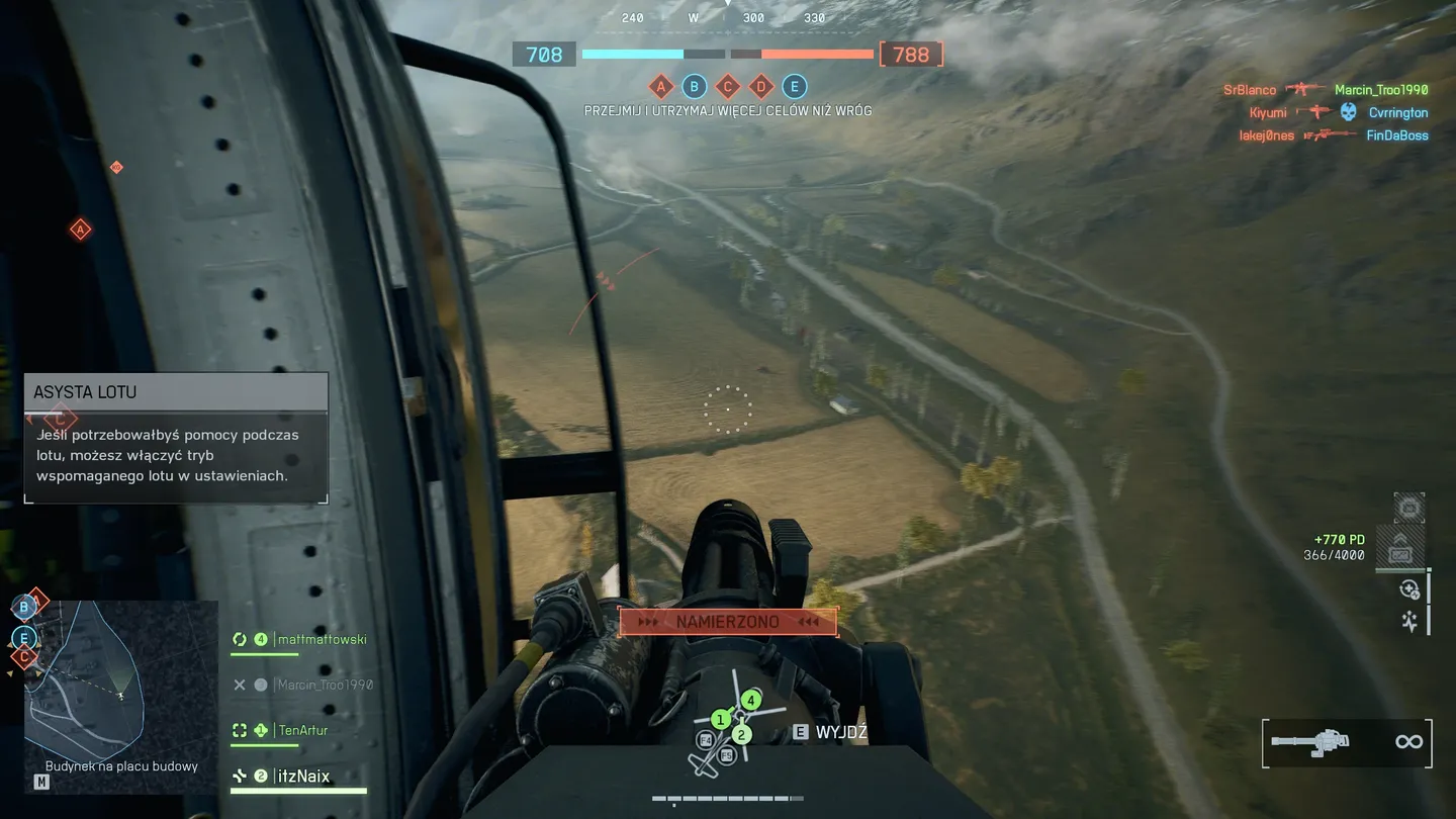 Widok z boku śmigłowca na otwarte tereny, pola i serpentynowe drogi &ndash; gracz kieruje ciężki karabin maszynowy, a na ekranie pojawia się informacja &bdquo;NAMIERZONO&rdquo; i instrukcja asysty lotu; w dolnym rogu interfejs pokazuje członk&oacute;w drużyny i mapę.