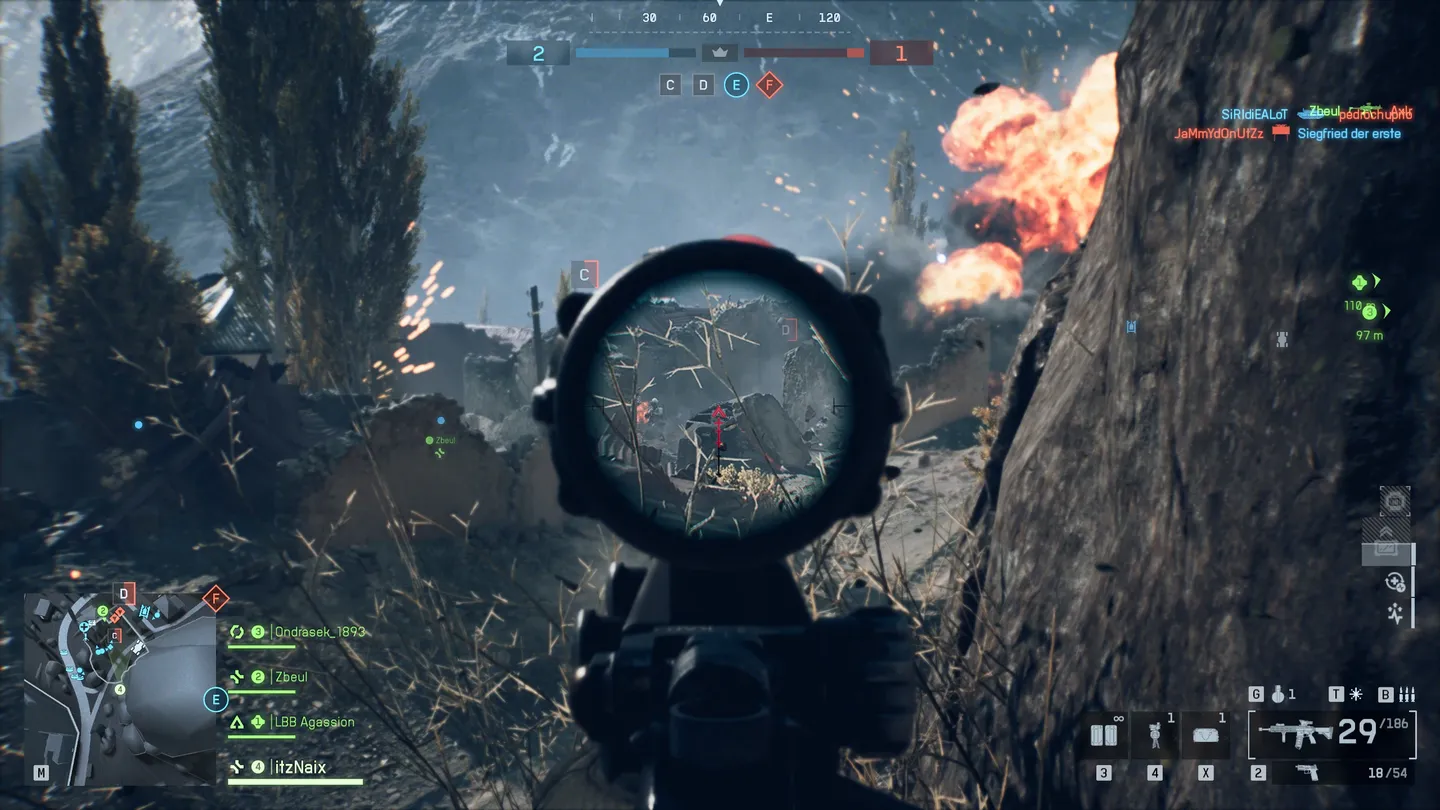 Widok przez celownik karabinu, skupiony na przeciwniku chowającym się za ruinami budynku, na tle wybuch&oacute;w; na dole ekranu widoczny jest HUD z mapą, status drużyny i licznik amunicji.