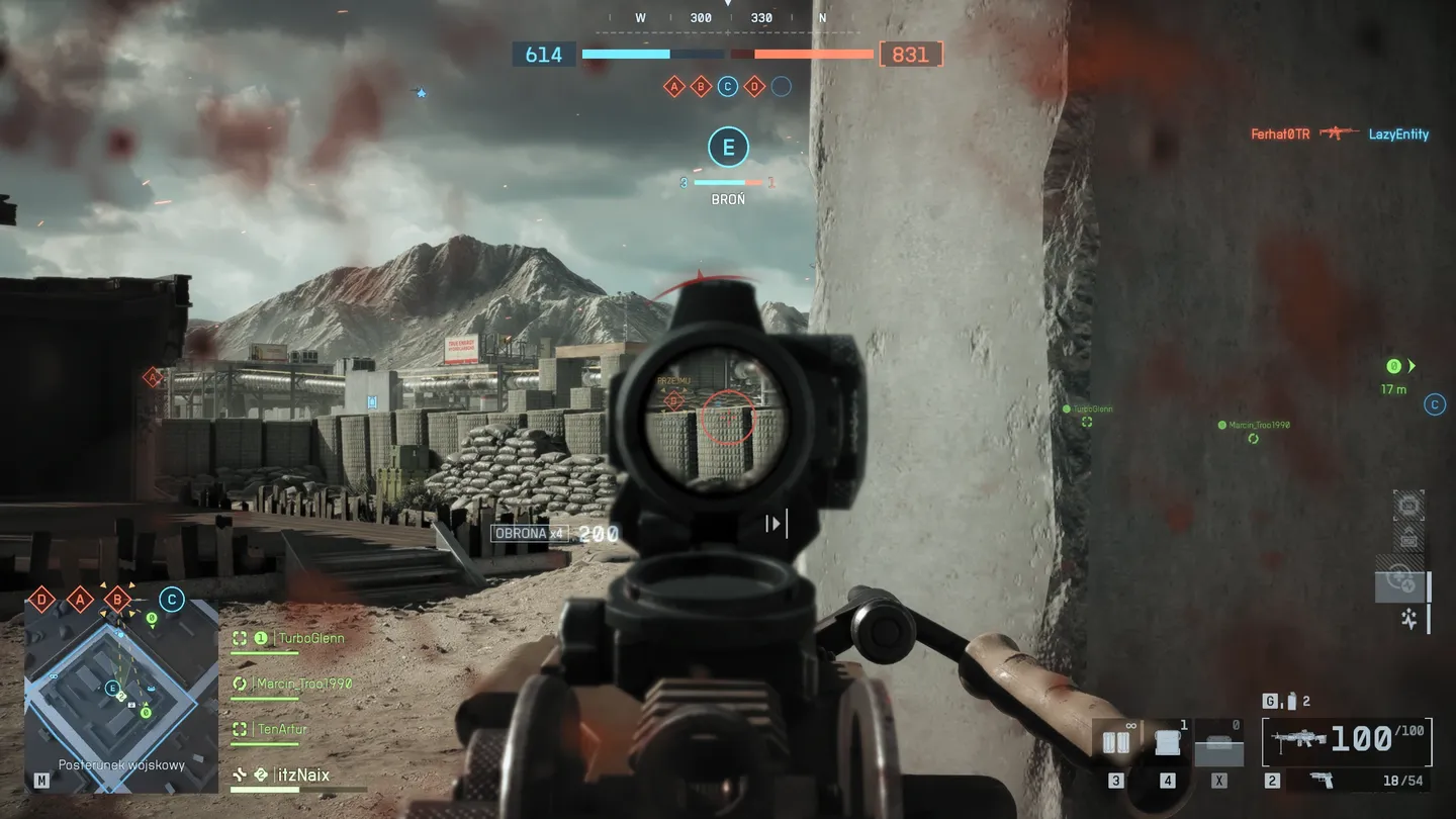 Rozgrywka na otwartej, częściowo zrujnowanej mapie &ndash; gracz przejmuje punkt A, trzymając ciężki karabin maszynowy, obok opancerzone pojazdy i sojusznicy; widoczne HUD, minimapa i licznik przejęcia punktu.