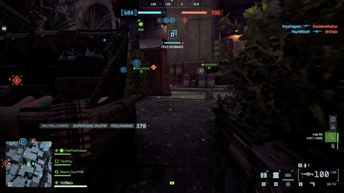 Widok z perspektywy pierwszej osoby podczas przejmowania punktu w ciasnej, zarośniętej alejce &ndash; po lewej stronie ekran wypełniają skrzynie i minimapa, na wprost widoczny budynek oraz oznaczenia kontrolowanych punkt&oacute;w.