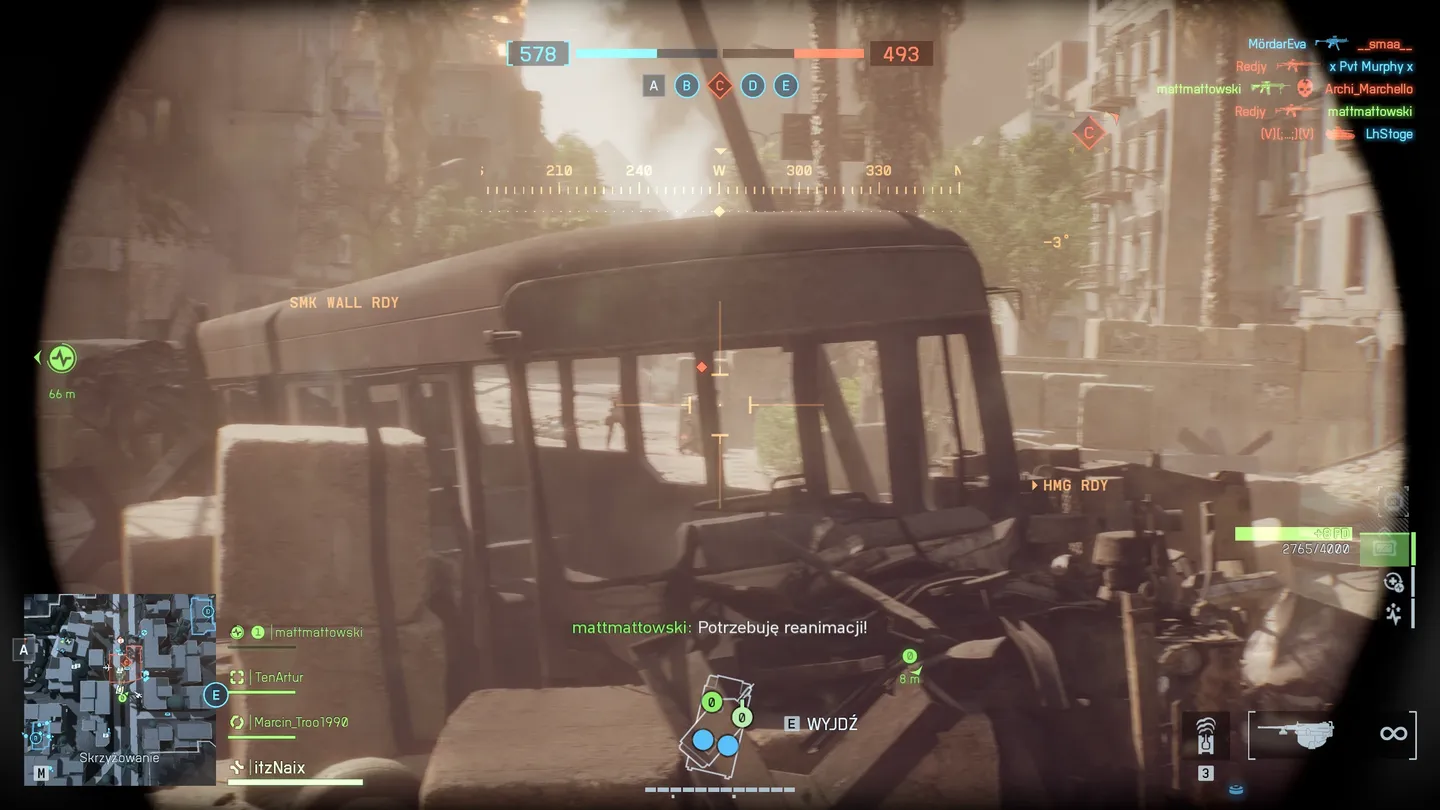 Widok z pojazdu opancerzonego na ulicę z wrakiem autobusu, barykadami i dymiącymi zgliszczami; na ekranie widoczne są oznaczenia pojazd&oacute;w, komunikat &bdquo;SMK WALL RDY&rdquo; oraz minimapa z pozycjami członk&oacute;w drużyny.
