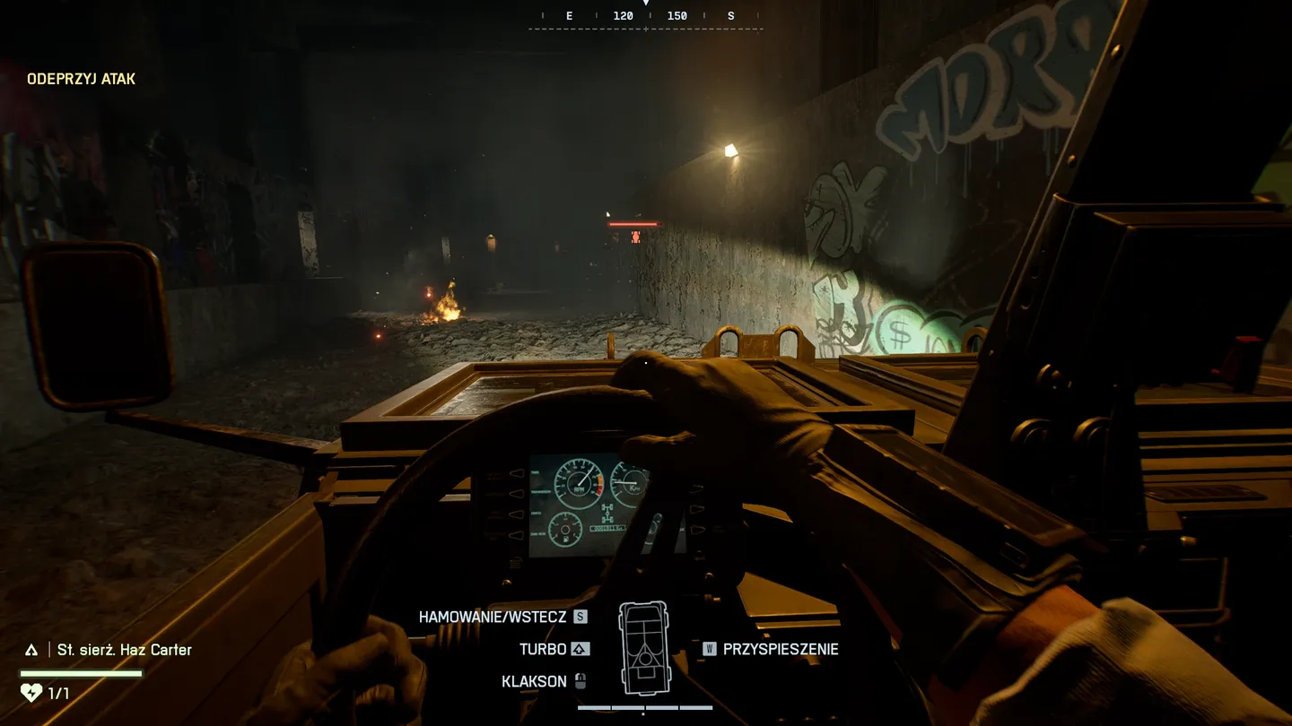 battlefield-6-singleplayer-recenzja-pograne-16.jpg: Widok z perspektywy pierwszej osoby podczas jazdy opancerzonym pojazdem przez zniszczony, ciemny tunel, na ścianach widoczne graffiti, na ziemi ogień i ruiny, a na ekranie wyświetlają się elementy interfejsu pojazdu oraz cele misji.