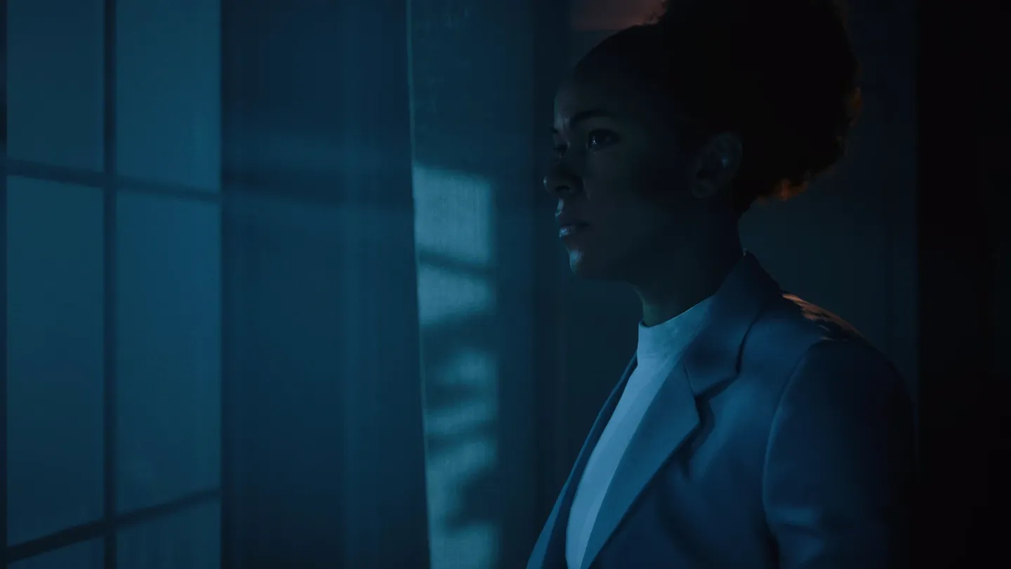 Dynamiczny portret kobiety w jasnym garniturze, stojącej przy oknie i patrzącej w dal w niebieskawym świetle wnętrza &ndash; scena sugeruje moment refleksji lub kluczową scenę fabularną.