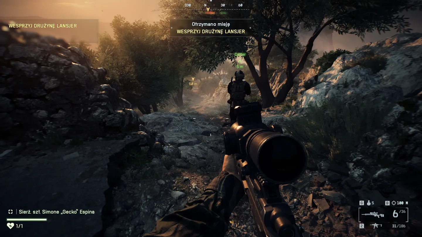 Ujęcie zza plec&oacute;w żołnierza patrolującego kamienistą, g&oacute;rską ścieżkę o zachodzie słońca, z widoczną bronią snajperską w dłoni gracza i zadaniem wspierania drużyny Lansjer.