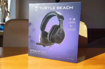 Alt: Pudełko słuchawek Turtle Beach Stealth 600 obok wyjętych z niego czarnych bezprzewodowych słuchawek z mikrofonem, na drewnianym stole w domowym otoczeniu.