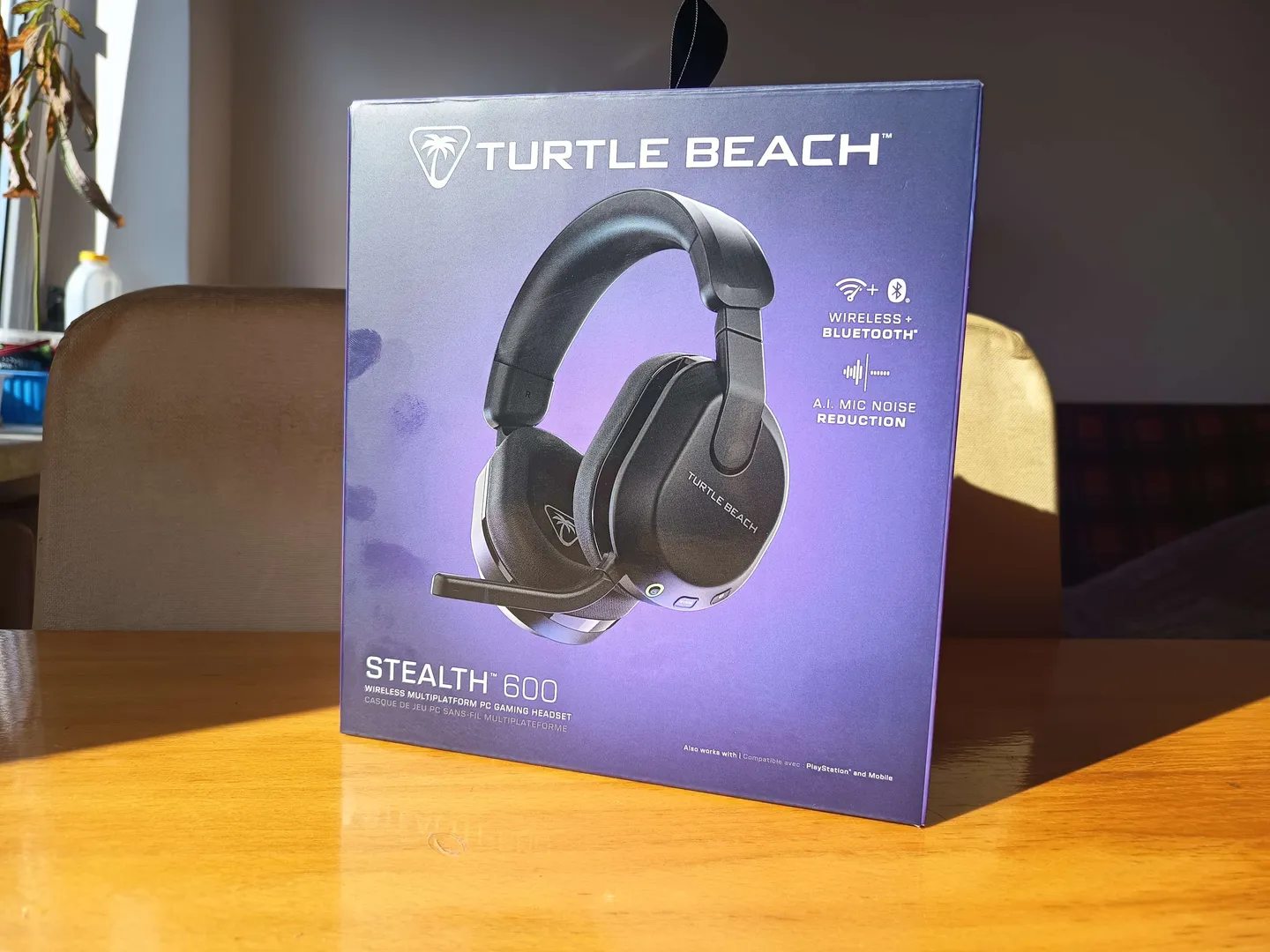 Alt: Pudełko słuchawek Turtle Beach Stealth 600 obok wyjętych z niego czarnych bezprzewodowych słuchawek z mikrofonem, na drewnianym stole w domowym otoczeniu.