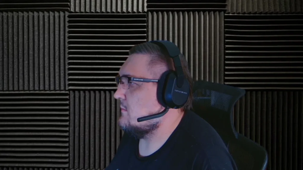 Profil mężczyzny z brodą i okularami, noszącego czarne bezprzewodowe słuchawki Turtle Beach Stealth 600 Gen 3 z mikrofonem, siedzącego w studiu z akustycznymi panelami i krzesłem gamingowym.