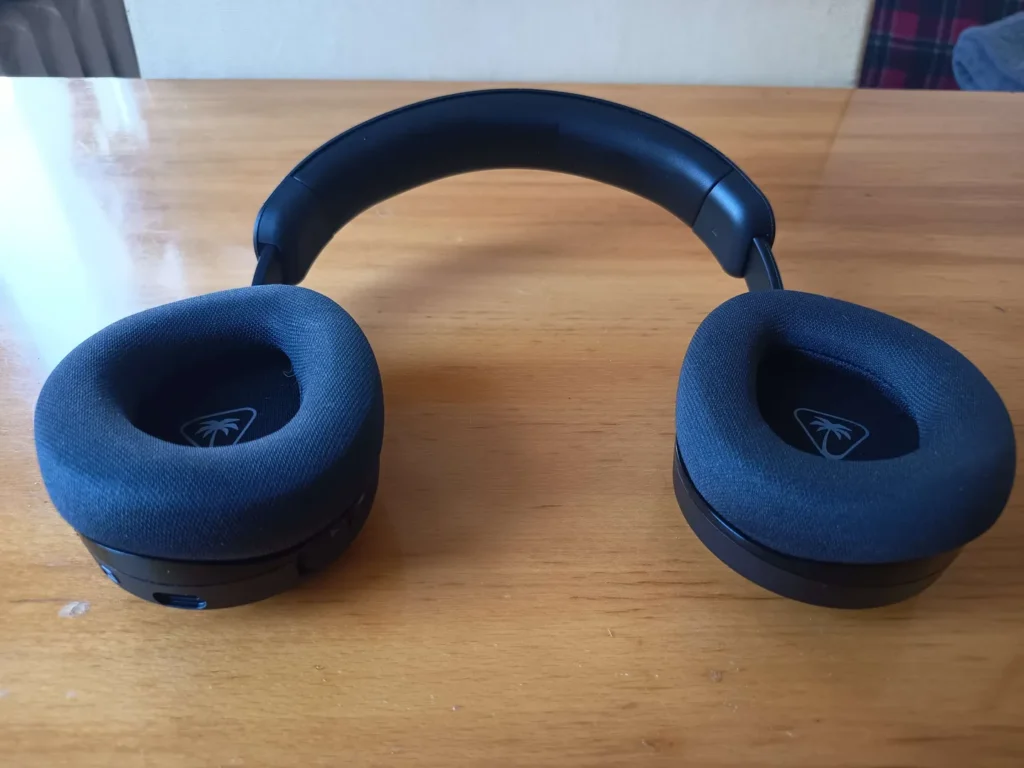 Widok z przodu słuchawek Turtle Beach Stealth 600 Gen 3 z miękkimi nausznikami z logo palmą na podkładkach i portem USB-C na spodzie, na drewnianym stole.