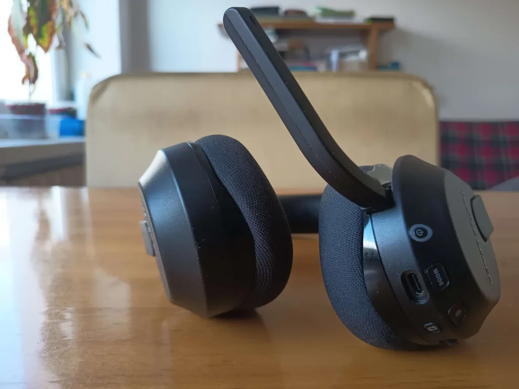 Zbliżenie na bok słuchawek Turtle Beach Stealth 600 Gen 3 z widocznym mikrofonem, portami USB-C i przyciskami na obudowie, stojących na drewnianym stole w domowym wnętrzu.