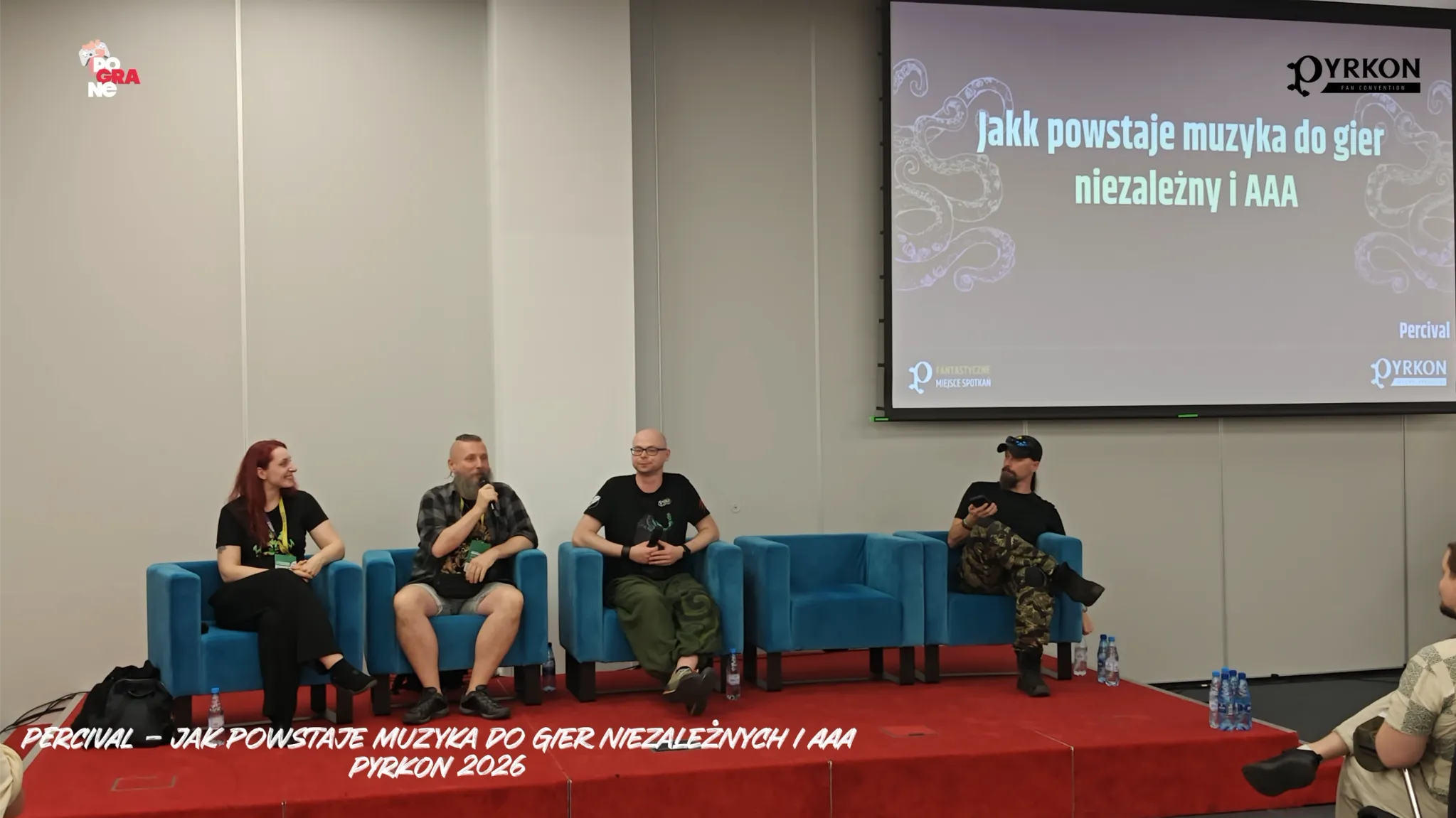 Zdjęcie przedstawia prelekcję zarejestrowaną podczas festiwalu Pyrkon 2026. Na dole kadru umieszczono biały napis na czerwonym tle: „PERCIVAL - JAK POWSTAJE MUZYKA DO GIER NIEZALEŻNYCH I AAA / PYRKON 2026”. W lewym górnym rogu widoczne jest również częściowo ucięte logo portalu Pograne.