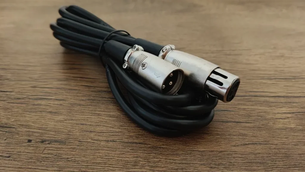 Zdjęcie przedstawia zwinięty, gruby przewód audio z interfejsem XLR, ułożony na jasnym, drewnianym blacie. Na pierwszym planie wyeksponowano dwie metalowe, srebrne wtyczki – trzybolcowe złącze męskie oraz pasujące złącze żeńskie z blokadą – zabezpieczone czarnymi, gumowymi odgiętkami chroniącymi kabel przed załamaniem.