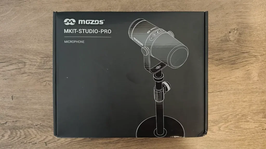 Zdjęcie przedstawia frontową stronę oficjalnego, matowego opakowania mikrofonu Mozos MKIT-STUDIO-PRO, sfotografowanego z góry na drewnianym podłożu. Minimalistyczna, czarna szata graficzna kartonu zawiera białe napisy z logo marki i oznaczeniem modelu po lewej stronie oraz dużą, białą grafikę wektorową przedstawiającą zarys mikrofonu na statywie.