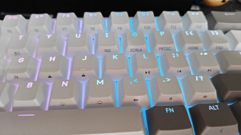 Zbliżenie na podświetlone klawisze białej klawiatury mechanicznej z niebiesko-fioletową iluminacją RGB i oznaczeniami funkcji bocznych.