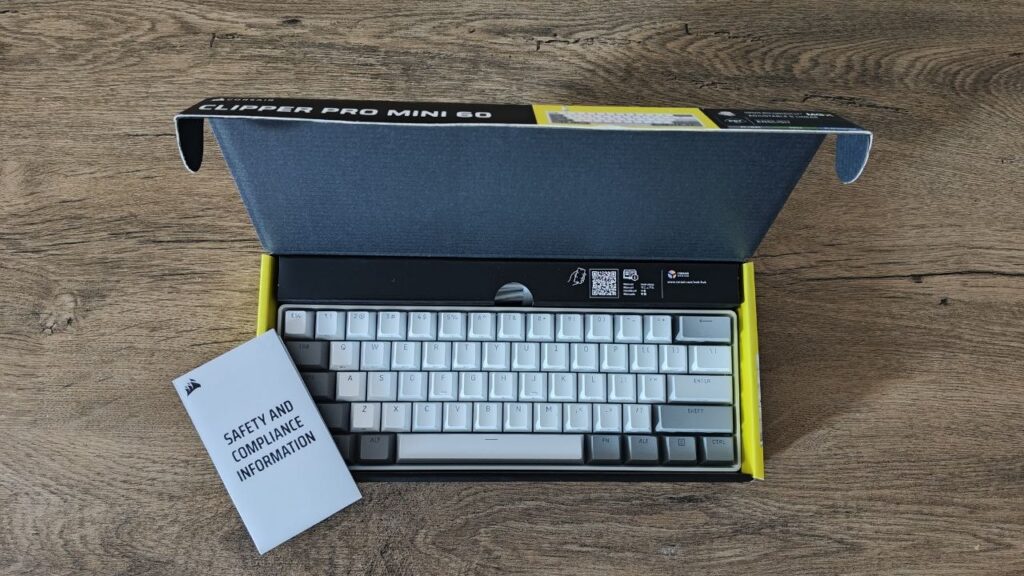 Otwarte żółto-czarne pudełko z klawiaturą Corsair Clipper Pro Mini 60 i dołączoną ulotką dotyczącą bezpieczeństwa.