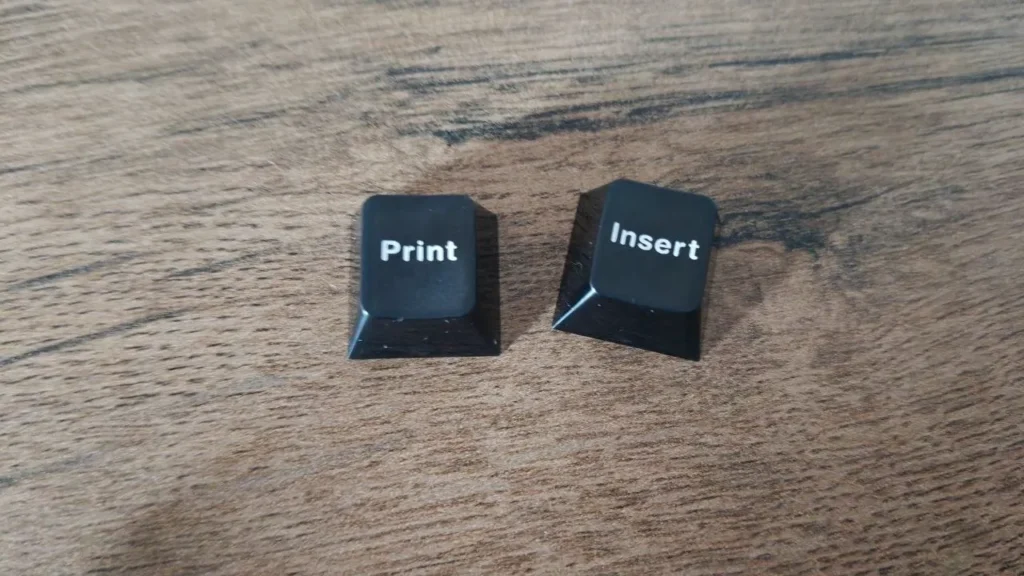 Dwa dodatkowe czarne keycapy z oznaczeniami Print oraz Insert leżące na blacie.