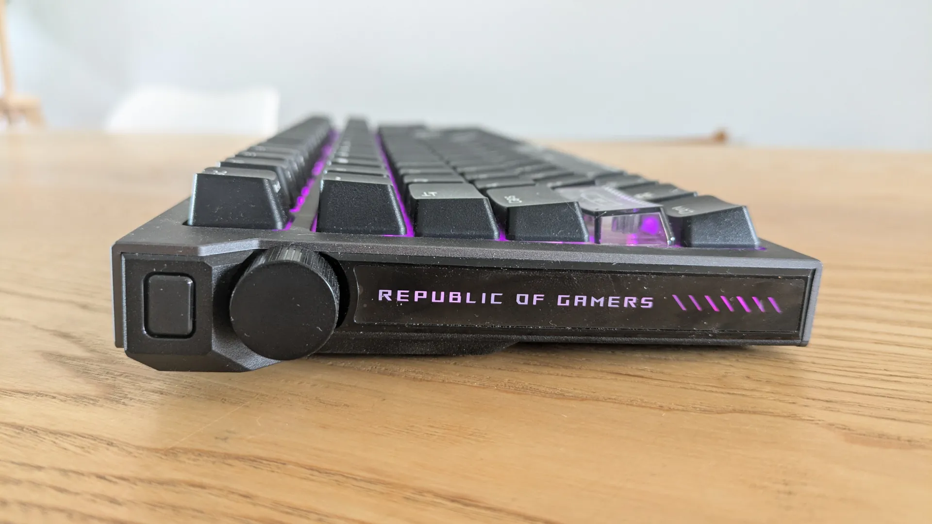 Bok klawiatury ASUS ROG Strix Morph 96 z widocznym napisem Republic of Gamers podświetlonym na fioletowo oraz okrągłym pokrętłem multimedialnym.