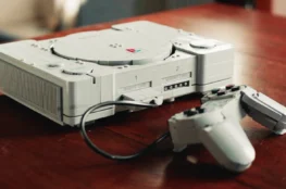 Zbudowana z jasnoszarych klocków LEGO replika klasycznej konsoli PlayStation 1 z podłączonym przewodowym kontrolerem i kartą pamięci na drewnianym stole.