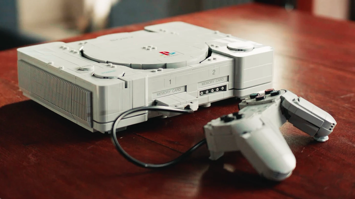 Zbudowana z jasnoszarych klocków LEGO replika klasycznej konsoli PlayStation 1 z podłączonym przewodowym kontrolerem i kartą pamięci na drewnianym stole.