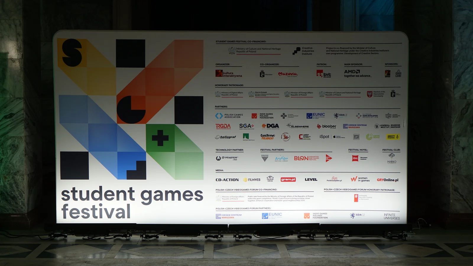 Na obrazie widoczny jest biały panel z logo i nazwą „student games festival”. Panel zawiera również logo i nazwy sponsorów i partnerów, w tym nazwy takich jak AMD, DGA, Intel, i innych.